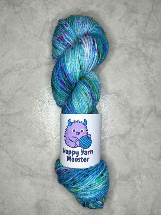 1 skein of hand-dyed yarn, on the DK Sock Monster base. In the colorway Ocean Party, which has a variegated blue base with yellow, green and pink speckles. The skein has a "Happy Yarn Monster" label.