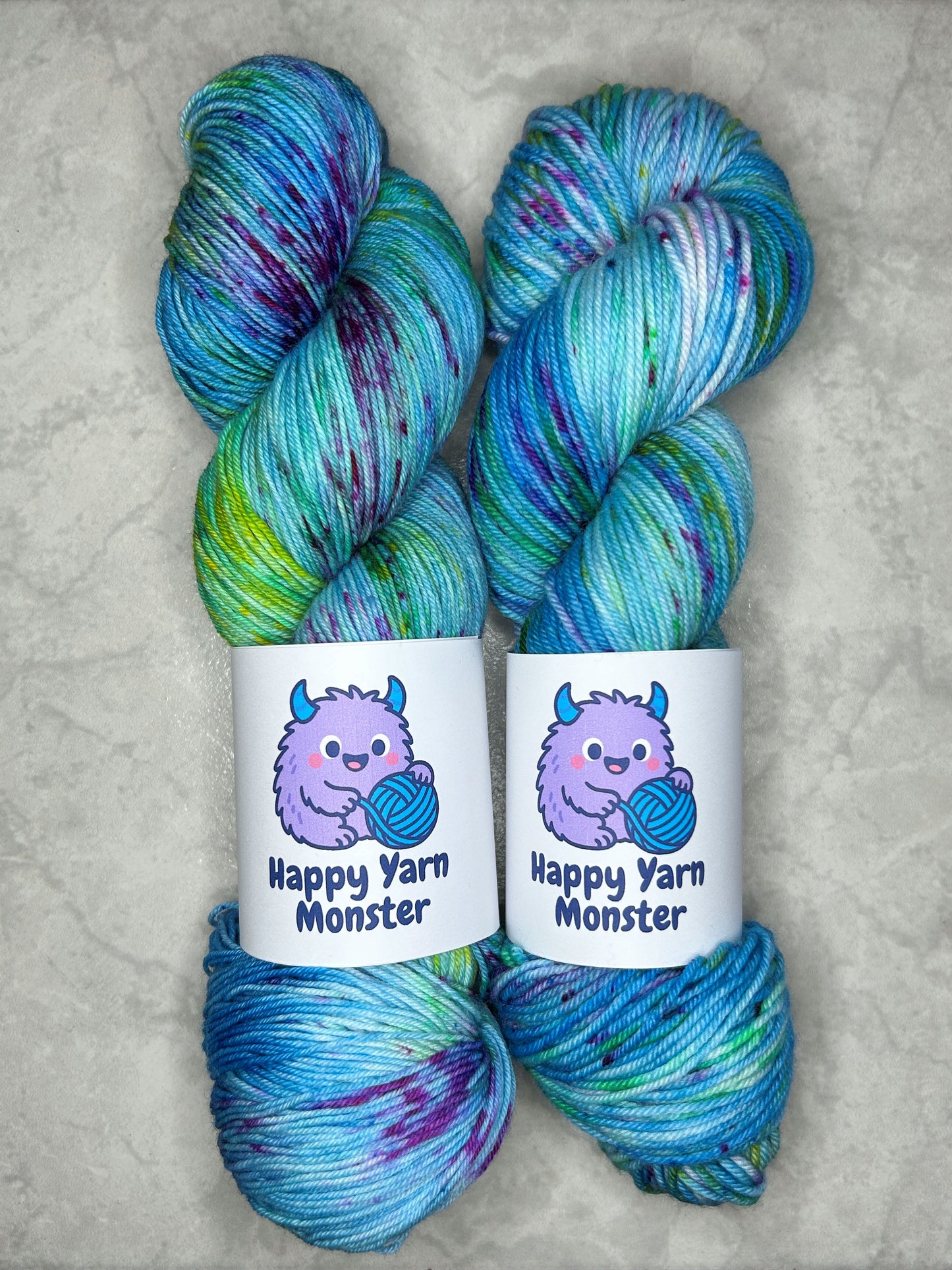2 skeins of hand-dyed yarn, both in the colorway Ocean Party, which have a variegated blue base with yellow, green and pink speckles. Both skeins have a "Happy Yarn Monster" label.