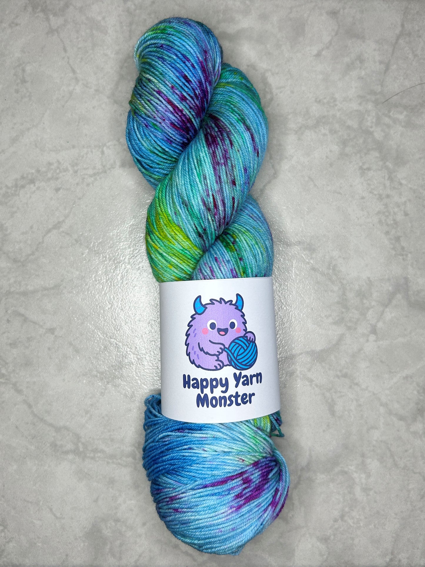 1 skein of hand-dyed yarn, on the Sock Monster base. In the colorway Ocean Party, which has a variegated blue base with yellow, green and pink speckles. The skein has a "Happy Yarn Monster" label.