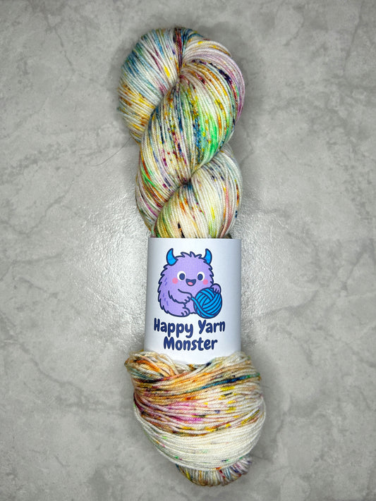 1 skein of hand-dyed yarn, on the Sock Monster base. In the colorway Rainbow Sprinkles, which has an undyed base with 7 rainbow colored peckles. The skein has a "Happy Yarn Monster" label.
