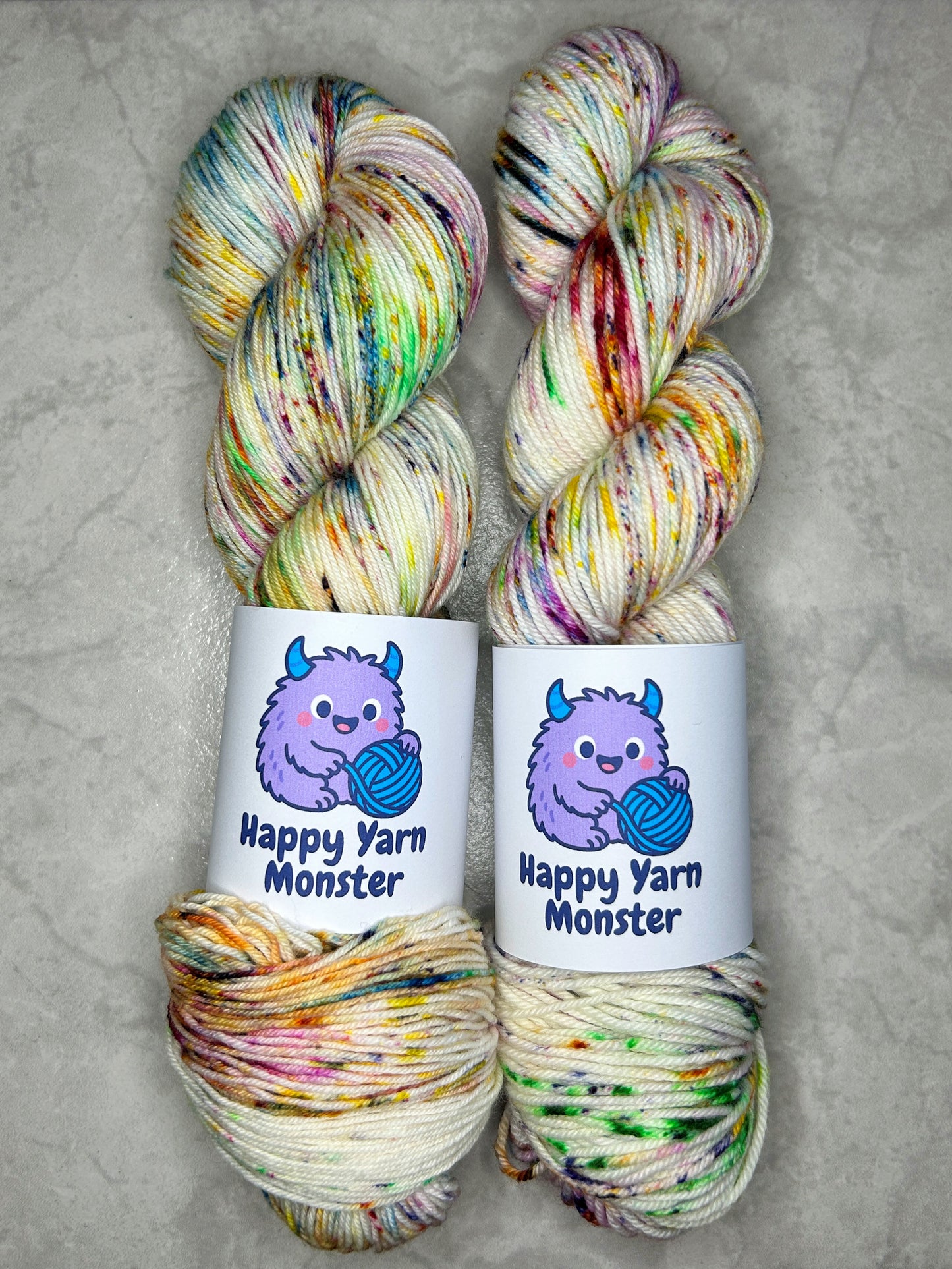 2 skeins of hand-dyed yarn, both in the colorway Rainbow Sprinkles, which have an undyed base with 7 rainbow colored peckles. Both skeins have a "Happy Yarn Monster" label.