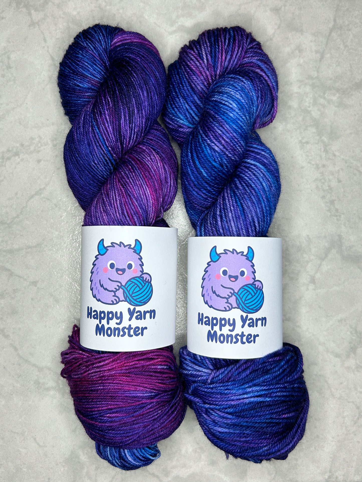 2 skeins of hand-dyed yarn, both in the colorway Fairy Night, which have variegated pink, blue en purples. Both skeins have a "Happy Yarn Monster" label.