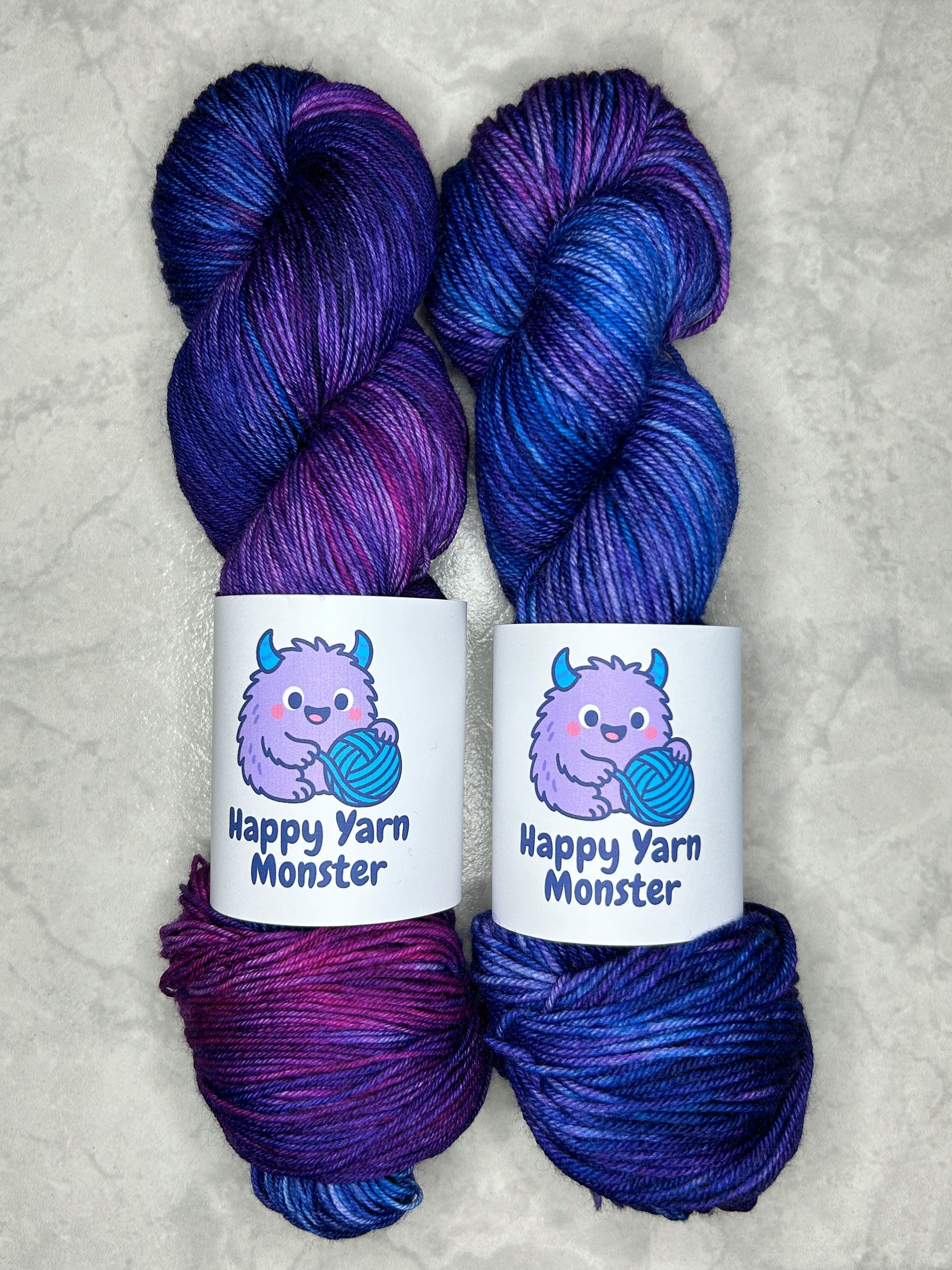 2 skeins of hand-dyed yarn, both in the colorway Fairy Night, which have variegated pink, blue en purples. Both skeins have a "Happy Yarn Monster" label.