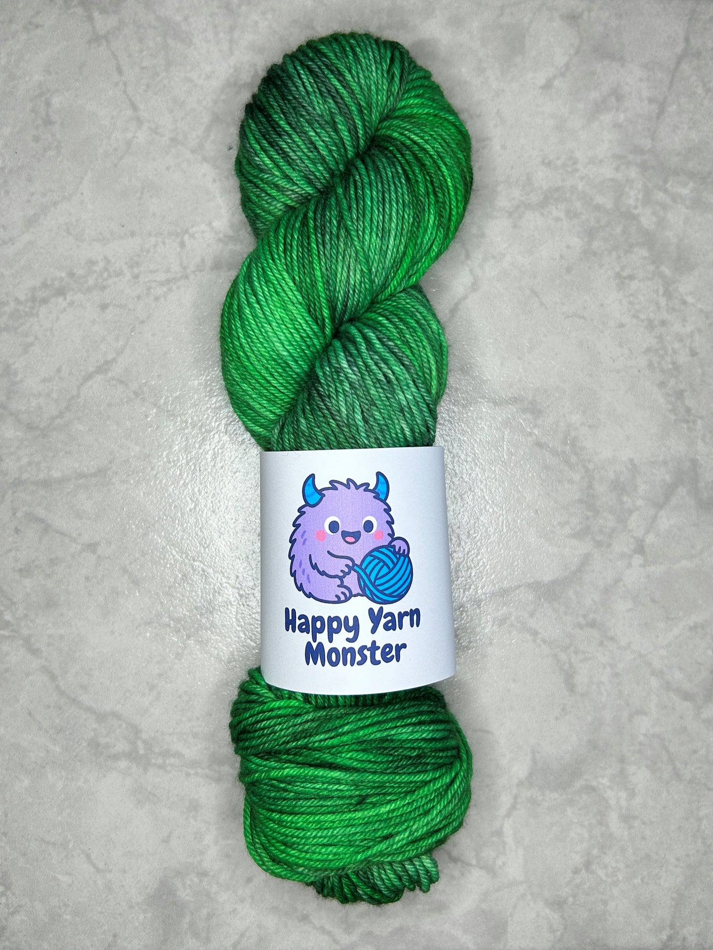 1 skein of hand-dyed yarn, on the DK Sock Monster base. In the colorway Forest Stroll, which has a green variegated base with darker and lighter greens. The skein has a "Happy Yarn Monster" label. 