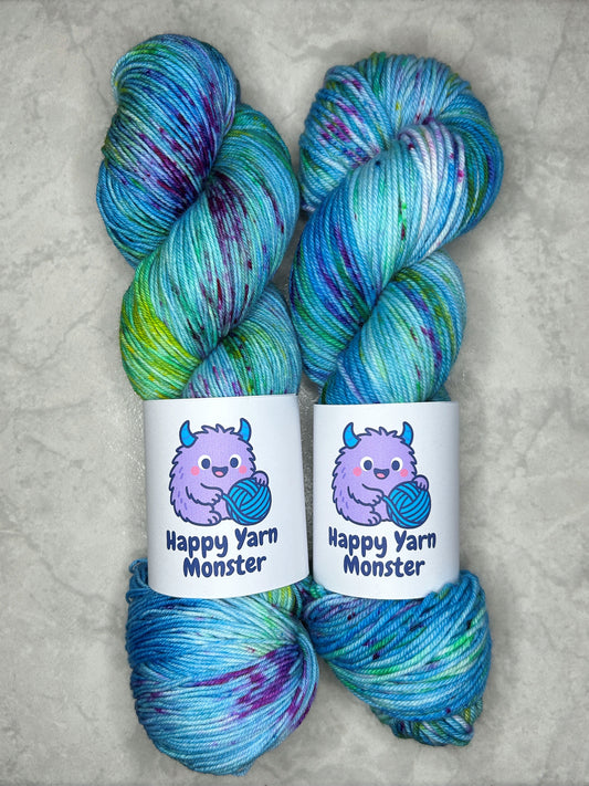 2 skeins of hand-dyed yarn, both in the colorway Ocean Party, which have a variegated blue base with yellow, green and pink speckles. Both skeins have a "Happy Yarn Monster" label.