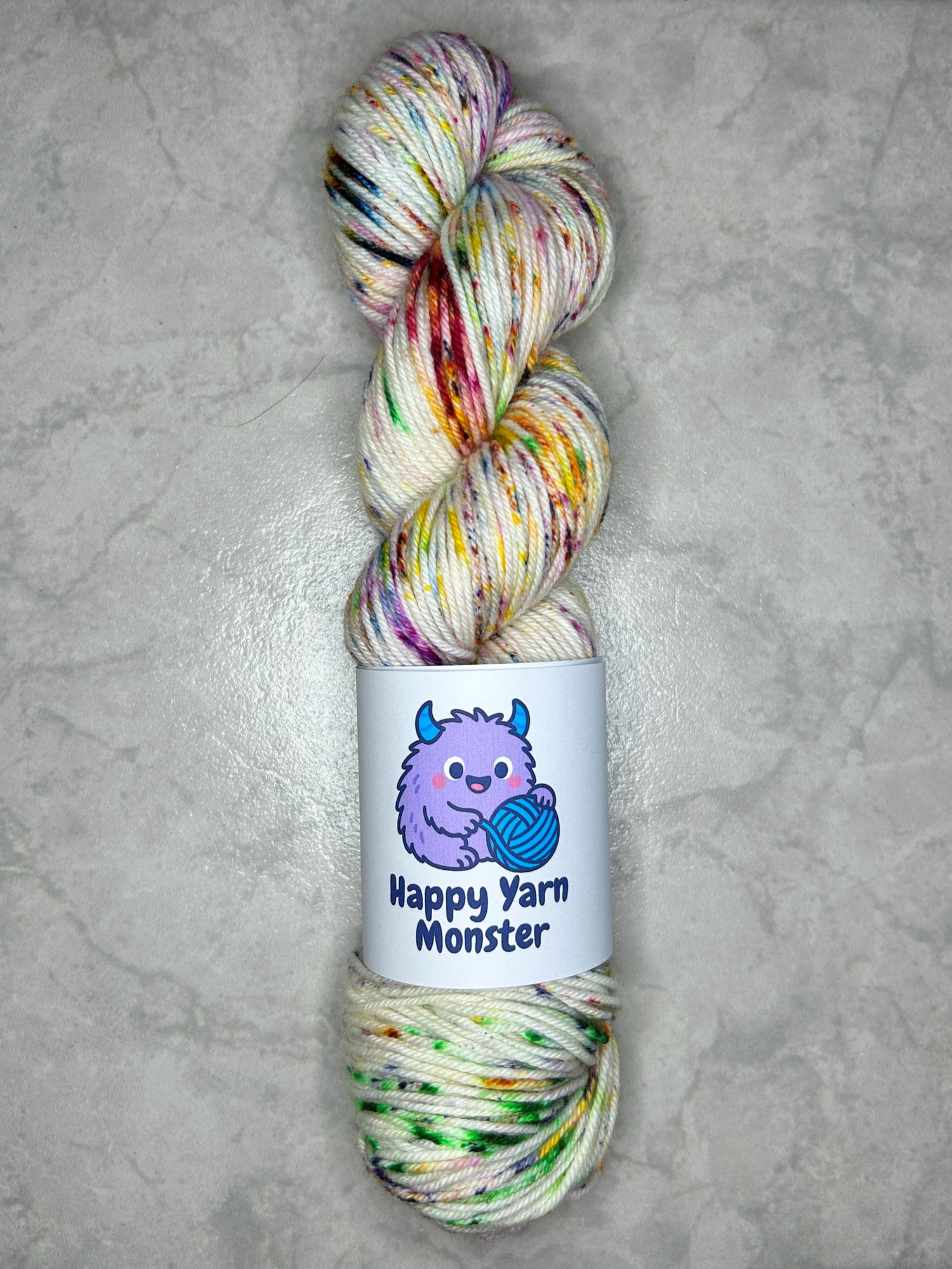 1 skeins of hand-dyed yarn, on the DK Sock Monster base. In the colorway Rainbow Sprinkles, which has an undyed base with 7 rainbow colored peckles. The skein has a "Happy Yarn Monster" label.